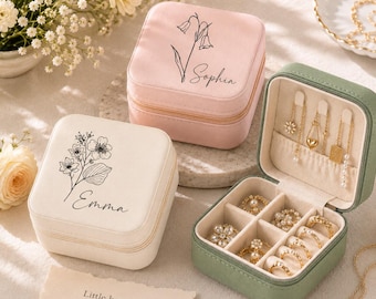 May include: Three square jewelry boxes in pink, white, and sage green. The pink and white boxes feature black floral line art and the names Sophia and Emma. The open green box contains gold necklaces and rings in velvet-lined compartments.