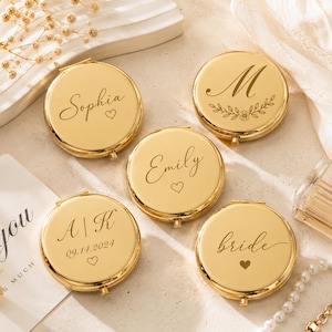 May include: Five round gold-coloured metal compact mirrors with personalised engravings. The mirrors feature various inscriptions including the names Sophia, Emily, the letter M with a floral wreath, the initials A | K with 09.14.2024, and the word bride.