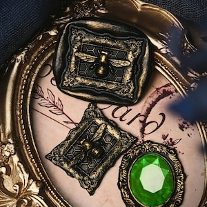 May include: Two rectangular wax seals featuring a gold bee centered on a honeycomb pattern within an ornate frame, and one oval wax seal with a faceted bright green gemstone, all resting inside a large, oval, gold-colored decorative frame.