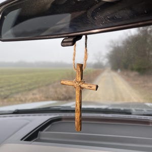 May include: A wooden cross hanging from the rearview mirror of a car. The cross is made of natural wood and has a simple design. The cross is hanging from a string made of natural fibers.