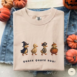 May include: A light beige t-shirt featuring a graphic of four ducklings in Halloween costumes: a witch, a mummy, a pirate, and a pumpkin. Below the ducks, the text reads QUACK QUACK BOO! in dark blue letters. The shirt has a crew neckline.