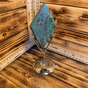 Peut inclure: Une pierre précieuse verte et violette de forme losange est exposée sur un support argenté. Le support a une base ronde avec un motif décoratif.