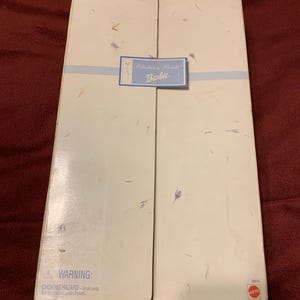 Peut inclure: Une boîte en carton blanche avec un ruban bleu et un autocollant qui dit "Blushing Bride Barbie". La boîte porte une étiquette d'avertissement qui dit "AVERTISSEMENT : RISQUE D'ÉTOUFFEMENT - Petites pièces. Ne convient pas aux enfants de moins de 3 ans."