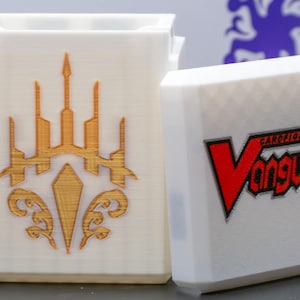 May include: A white rectangular card storage box with a detachable lid. The front of the box features a gold-colored emblem, while the lid displays the red and black Cardfight!! Vanguard logo. The surface has a visible 3D printed texture.