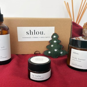 May include: A collection of skincare products, including body oil, body butter, and lip balm, arranged on a red surface. The products are in amber glass bottles and jars with white labels. A small felt Christmas tree and a cardboard box are in the background.