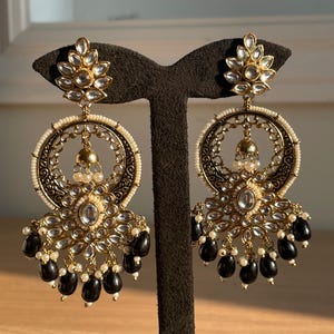 May include: A pair of gold earrings with black and white beads. The earrings have a circular design with intricate details and a floral motif at the top.