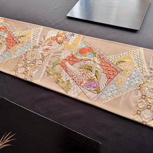 May include: A beige decorative table runner with embroidered patches featuring floral and bird designs in red, gold, and blue. The runner has a rectangular shape and is placed on a black surface.