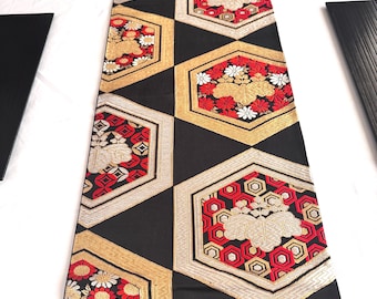 May include: A decorative table runner with a geometric pattern. The runner features black, gold, red, and white floral and geometric designs. The pattern is composed of hexagonal shapes with floral and geometric motifs.