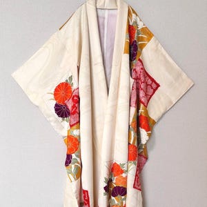 May include: A cream-colored kimono featuring a subtle tonal wave pattern and large, vibrant floral panels. The design includes bright orange, deep purple, and red chrysanthemum-like flowers, along with intricate red and white geometric patterns.
