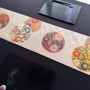May include: Cream-colored table runner with a repeating pattern of colorful floral medallions. The medallions feature intricate designs in red, yellow, blue, and white. Two black serving trays are also visible.