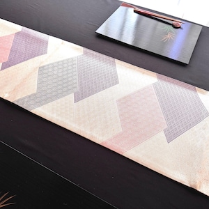 May include: A decorative table runner with a geometric pattern in shades of pink, gray, and cream. The runner is placed on a dark surface, with a black tray and chopsticks visible in the background. The runner's design features repeating shapes.
