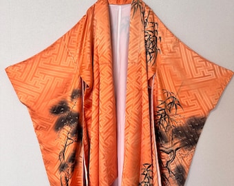 May include: An orange kimono featuring a tonal geometric pattern and detailed black ink-style illustrations of pine trees and bamboo stalks. The garment has wide sleeves, a structured collar, and a light-colored interior lining, hanging on a metal hanger.