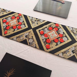 以下が含まれることがあります： 金、赤、銀色の花の刺繍が施された黒いテーブルランナー。 ランナーは正方形とひし形の幾何学模様のデザインです。 背景は明るい色の表面です。