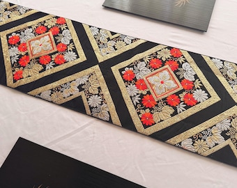 May include: Black table runner with a repeating pattern of gold, red, and silver floral embroidery. The runner features a geometric design with square and diamond shapes. The background is a light-colored surface.