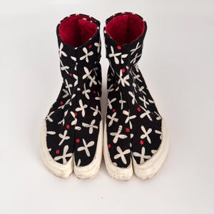 May include: A pair of black high-top tabi boots featuring a white cross-shaped pattern and small red dots. The boots have a split-toe design, white rubber soles, and a solid red interior lining visible at the top opening of each boot.