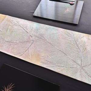 May include: A rectangular, iridescent table runner with a textured, white, fern-like pattern. Two black serving trays are visible, one with a similar fern design and chopsticks. The runner has a soft, pastel color palette.