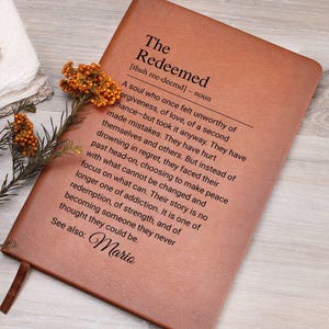 May include: A brown leather journal with the word "The Redeemed" embossed on the front cover. The text defines the word "Redeemed" as a soul who has overcome adversity and found peace. The name "Mario" is written at the bottom of the page.