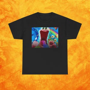 Op de afbeelding: Zwart T-shirt met een kleurrijke afbeelding. De afbeelding toont een fantasieachtig wezen met grote ogen, een rode lichaamskleur, een regenboog, bloemen en andere fantasie-elementen. Het T-shirt is gemaakt van een zacht materiaal.