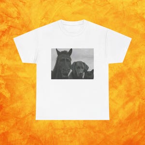 Puede incluir: Camiseta blanca con una imagen en escala de grises de un caballo, un perro y un gato. La imagen está centrada en la parte delantera de la camiseta. La camiseta tiene cuello redondo y mangas cortas. El fondo es naranja.