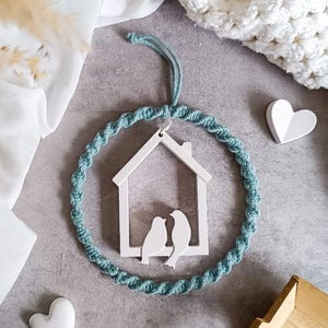 Könnte beinhalten: Ein weißes, hausförmiges Ornament aus Holz mit zwei Vogelsilhouetten in einem türkisfarbenen Makramee-Kreis. Das Ornament hängt an einer türkisfarbenen Schnur. Zwei herzförmige Dekorationen in Weiß sind ebenfalls zu sehen.