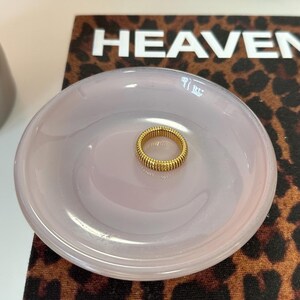 Könnte beinhalten: Ein goldfarbener Ring mit geripptem Design liegt in einer hellrosa, runden Schale. Die Schale befindet sich auf einer Oberfläche mit Leopardenmuster und dem Wort "HEAVEN" in weißen Blockbuchstaben.