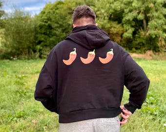 May include: Black hoodie with three orange chili pepper graphics on the back. The hoodie is worn by a person in a grassy field with trees in the background. The person is wearing gray sweatpants.