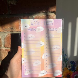 May include: A pastel-coloured weekly lists notepad. The notepad has sections for "MUST DO" and "COULD DO" with the text "WEEKLY LISTS" at the top. The design includes cute cloud illustrations. The background has a gradient of pink, yellow and orange.