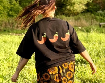 May include: Black t-shirt featuring three orange chilli pepper graphics. The person is wearing a sunflower print skirt. The background is a green field with trees.