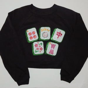 Puede incluir: Sudadera corta negra con cinco fichas de Mahjong con lentejuelas. Las fichas presentan círculos rojos, una ficha de "Joker" y caracteres chinos. Las fichas están bordeadas con lentejuelas verdes.