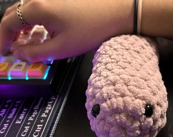 May include: A light pink, crocheted, plush snake toy with two small, round, black plastic eyes. The plush snake is positioned next to a black mechanical keyboard with white and yellow keycaps that feature various icons and symbols.