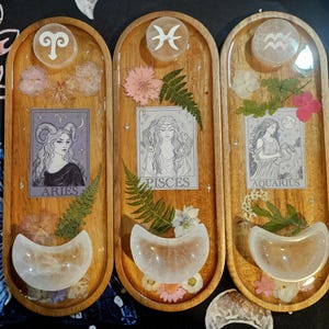 May include: Three wooden trays with resin and botanical elements. Each tray features a zodiac sign, a portrait of a woman, dried flowers, and a clear, crescent-shaped stone. The signs are Aries, Pisces, and Aquarius. The trays are oval-shaped.