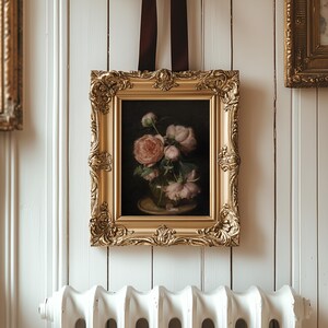 May include: A framed floral still life painting with a gold ornate frame. The painting depicts pink roses in a glass vase. The frame is suspended by a dark brown ribbon against a white paneled wall.