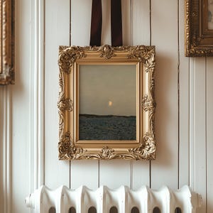 May include: A framed landscape painting with a gold ornate frame is suspended by a dark brown ribbon. The painting depicts a seascape with a moon or sun in the sky. The frame is rectangular with detailed carvings.