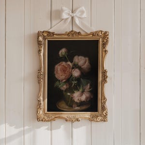 May include: A framed floral still life painting featuring pink peonies in a glass vase. The painting is set against a dark background and is enclosed in an ornate gold frame. A white ribbon is tied at the top for hanging.
