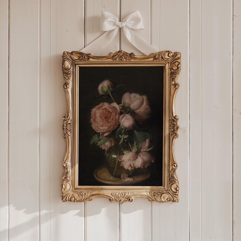 May include: A framed floral still life painting featuring pink peonies in a glass vase. The painting is set against a dark background and is enclosed in an ornate gold frame. A white ribbon is tied at the top for hanging.