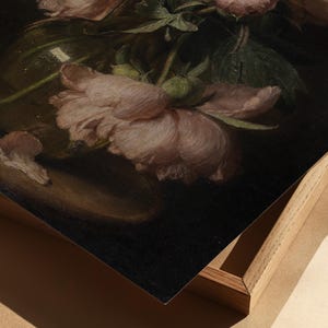May include: A still life painting featuring soft pink peonies in a clear glass vase. The flowers are in full bloom, with delicate petals and green leaves. The painting is framed in a light wood frame.