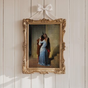 Pu&ograve; includere: Un dipinto incorniciato raffigurante una scena romantica di un uomo e una donna che si abbracciano. L'ornata cornice dorata &egrave; sospesa da un nastro bianco legato a fiocco. Il dipinto mostra una donna in abito blu e un uomo in cappotto marrone.