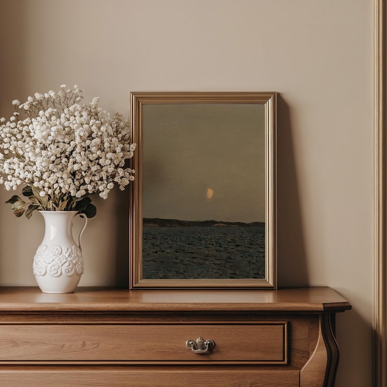 May include: A framed landscape painting of the sea and sky with a golden frame. A white vase with white flowers sits to the left of the painting on a wooden dresser. The painting depicts a seascape with a crescent moon.
