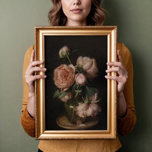 May include: A framed floral still life painting featuring pink peonies in a glass vase. The artwork has a dark background and is set in a gold ornate frame. The painting evokes a vintage aesthetic, with soft lighting and delicate brushstrokes.