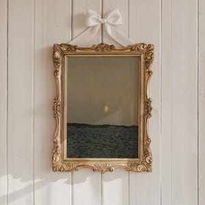 May include: A framed landscape painting depicting a seascape under a dusky sky with a crescent moon. The ornate gold frame is suspended by a white ribbon tied in a bow. The artwork features shades of blue, gray, and gold.