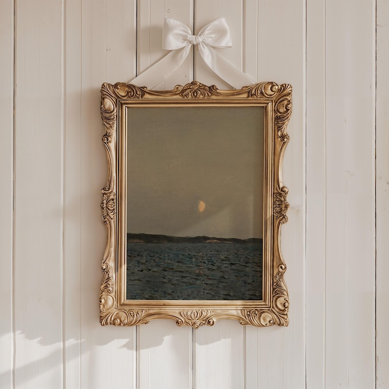 May include: A framed landscape painting depicting a seascape under a dusky sky with a crescent moon. The ornate gold frame is suspended by a white ribbon tied in a bow. The artwork features shades of blue, gray, and gold.
