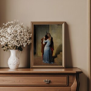 Pu&ograve; includere: Una stampa incorniciata di un dipinto classico raffigurante una coppia che si abbraccia. La donna indossa un abito blu e l'uomo un cappotto marrone. L'opera d'arte &egrave; in una cornice dorata, esposta su una cassettiera in legno con un vaso bianco di fiori.