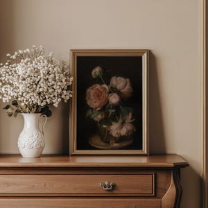 May include: A still life featuring a framed painting of pink peonies in a glass vase, set on a wooden dresser. A white ceramic pitcher with a floral design holds a bouquet of white flowers. The painting has a gold frame.