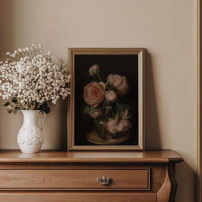 May include: A still life featuring a framed painting of pink peonies in a glass vase, set on a wooden dresser. A white ceramic pitcher with a floral design holds a bouquet of white flowers. The painting has a gold frame.