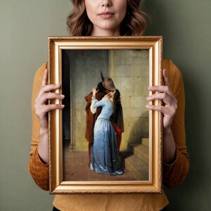 Pu&ograve; includere: Una stampa incorniciata di un dipinto classico raffigurante una scena romantica. L'opera presenta un uomo con un cappotto e un cappello marrone che abbraccia una donna con un vestito blu. La cornice &egrave; dorata e lo sfondo mostra un muro di pietra e delle scale.
