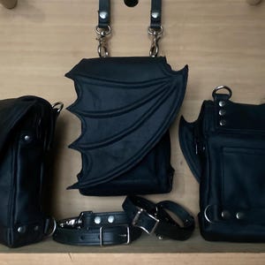 May include: Three black bags with bat wing-shaped flaps and silver-toned metal hardware, including two adjustable straps with buckles and rivets. The bags feature structured bodies and are displayed together with matching black leather-like straps.