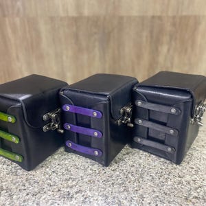 May include: Three black leather boxes with hinged lids and metal latches. Each box features three horizontal straps on the front, colored lime green, purple, and dark gray respectively, secured with silver-toned rivets on each side.