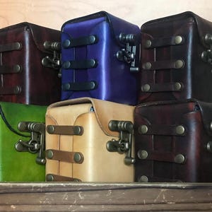May include: A collection of six square leather boxes in various colors including dark brown, purple, green, and tan. Each box features a flap closure secured with a metallic latch and decorative horizontal leather straps with brass-toned rivets.