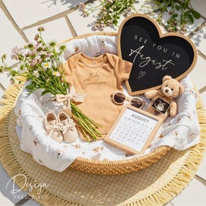 May include: A wicker basket holds baby items, including a tan onesie with "Baby Swanson" script, sunglasses, and a teddy bear holding an ultrasound image. A heart-shaped chalkboard reads "See you in August." A calendar shows the month of August.