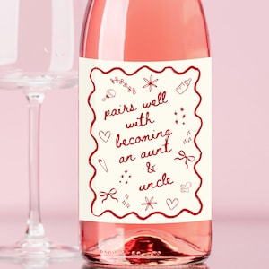 以下が含まれることがあります： 「pairs well with becoming an aunt & uncle」と書かれたラベルが付いたロゼワインのボトル。ラベルはクリーム色で、赤ちゃん関連のアイテムの赤いイラストが描かれています。背景にはワイングラスがあります。